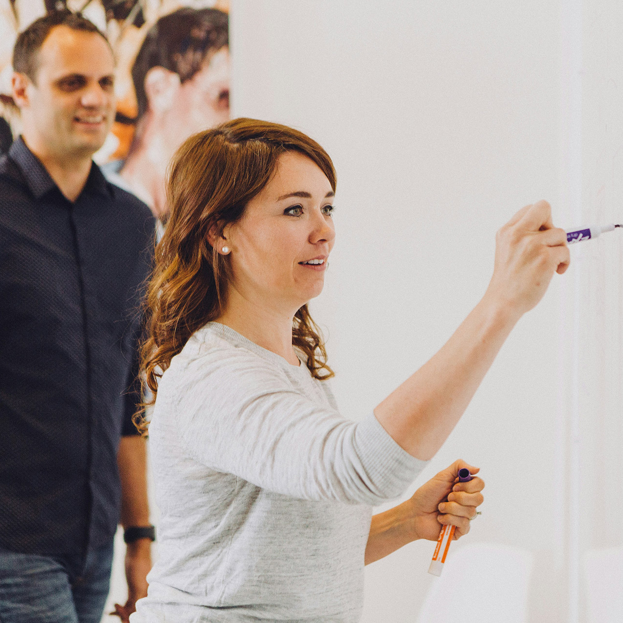 A woman with long red hair draws with a marker pen on a whiteboard or wall. A smiling man looks on from the background