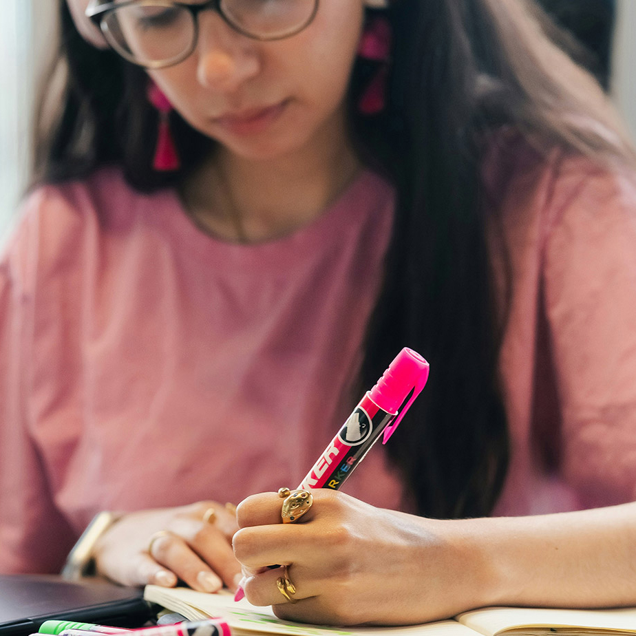 Close up of a young woman using a pink marker pen. The pen and her hand are in focus, her torso and face are blurred. SHe is wearing gold rings, a pink t-shirt and pink earrings, and has dark framed glasses on