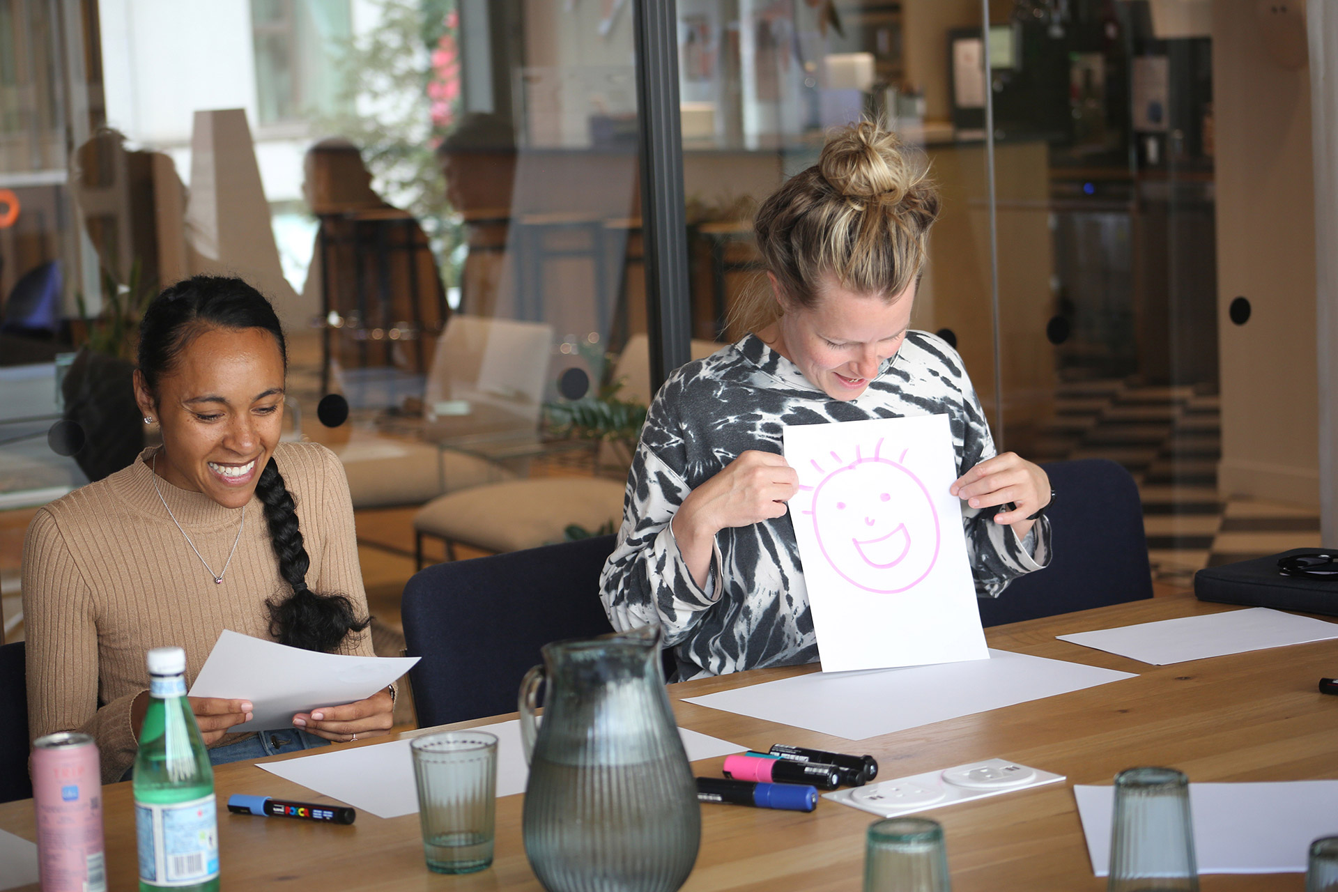 A woman smiles as she holds up her drawing of a smiley face, the young woman next to her is also smiling and holding a piece of paper. There are pens and a water jug and glasses on the table